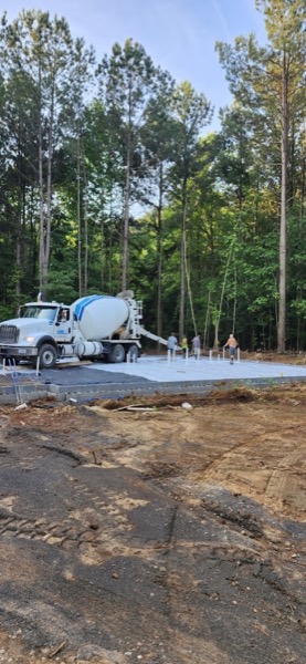 A concrete truck and crew screeding a fresh slab pour on a wooded lot